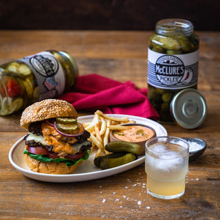 Burger with fries and a drink on a wooden table with McClure's Pickles in the background.